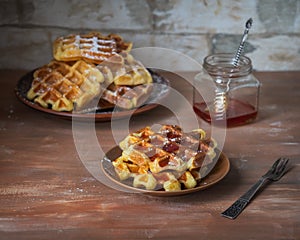 Sweet breakfast with waffles sprinkled with icing sugar on a small plate, a glass jar with fruit syrup and a dessert fork on a