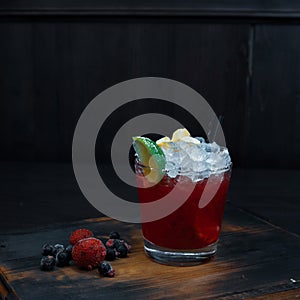 Sweet cold alcoholic cocktail of red color with fresh berries and lime slices stands on a wooden table in a pub.