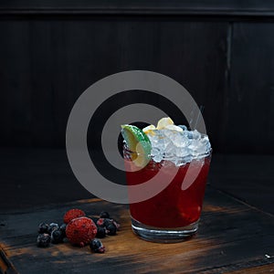 Sweet cold alcoholic cocktail of red color with fresh berries and lime slices stands on a wooden table in a pub. Cocktail served