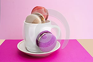 Sweet colorful macarons in white cup on pink background. Tasty red, green, brown, violet macaroons