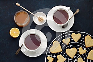 Sweet cookies in heart shape with two cups of tea, honey and lemon on dark blue background Valentines day.