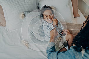 Sweet excited mixed race boy lying on bed and laughing while playing with his loving mom