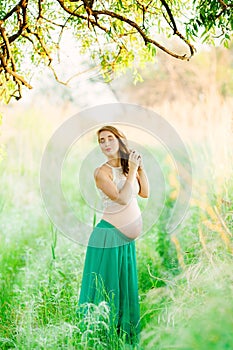 A sweet, fair-haired pregnant girl in a green skirt is standing in the park with her eyes closed and braids her braid