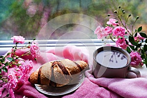 Sweet fresh morning coffee and croissant staying on the windowsill with pink flowers