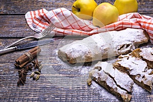 Sweet freshly baked homemade apple strudel with sugar powder and raw yellow apples on a wooden background.