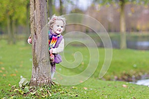 Sweet funny toddler girl hiding behind tree in park