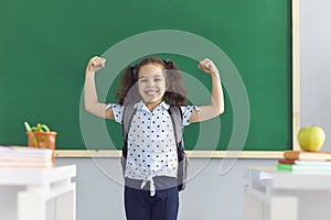 Sweet girl showing her muscles in classroom. Strong kid expressing motivation to achieve success. Back to school