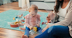 Sweet girl is sitting on carpet among toys with mother, holding bottle, cup in hands,
