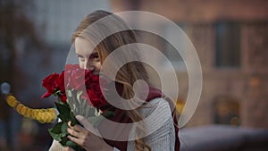 Sweet girl smelling bouquet flowers outside. Cute woman carrying roses on street