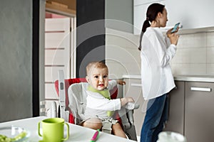 Sweet little baby in green bib sitting in baby chair getting scared seeing bird outside the window while mom turns aside