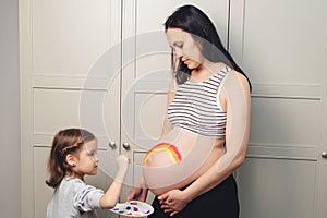 Sweet little daugter painting pregnant belly her mother. Baby birth expecting time and belly painting