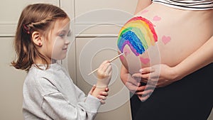 Sweet little daugter painting pregnant belly her mother. Pregnant mom and her child having fun together at home. Family, healthy