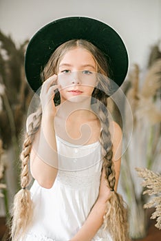 A sweet little girl with long blond hair in a white sarafan and a black hat in a rattan chair