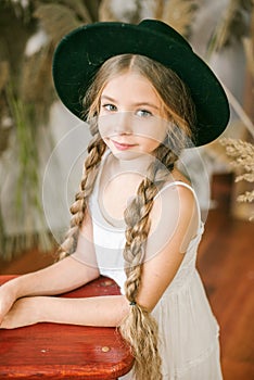 A sweet little girl with long blond hair in a white sarafan and a black hat in a rattan chair