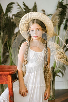 A sweet little girl with long blond hair in a white sarafan and a black hat in a rattan chair