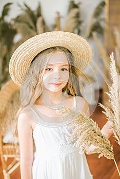 A sweet little girl with long blond hair in a white sarafan and a straw hat in a rattan chair