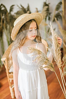 A sweet little girl with long blond hair in a white sarafan and a straw hat in a rattan chair