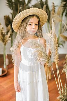 A sweet little girl with long blond hair in a white sarafan and a straw hat in a rattan chair