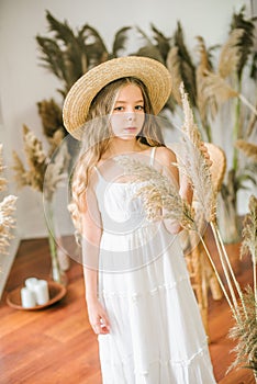 A sweet little girl with long blond hair in a white sarafan and a straw hat in a rattan chair