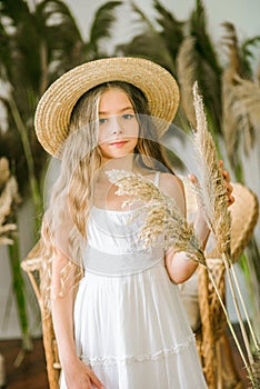 A sweet little girl with long blond hair in a white sarafan and a straw hat in a rattan chair