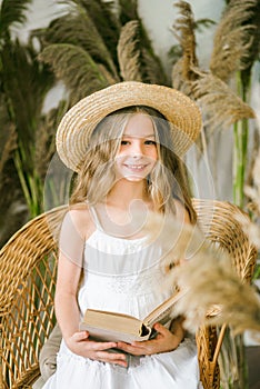 A sweet little girl with long blond hair in a white sarafan and a straw hat in a rattan chair