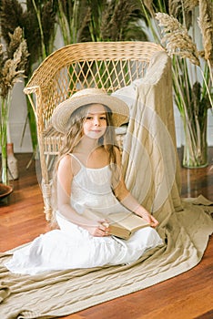 A sweet little girl with long blond hair in a white sarafan and a straw hat in a rattan chair