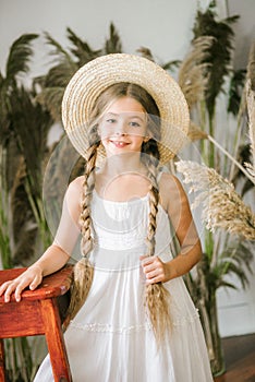 A sweet little girl with long blond hair in a white sarafan and a straw hat in a rattan chair