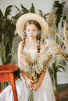 A sweet little girl with long blond hair in a white sarafan and a straw hat in a rattan chair