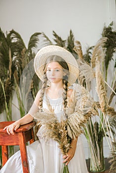 A sweet little girl with long blond hair in a white sarafan and a straw hat in a rattan chair