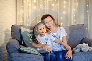 Sweet little girl together with beautiful young mom sitting on sofa in living room at home in evening. Happy family concept. Love