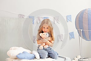 Sweet little redhead girl is hugging teddy bear on at home. Smiling little blonde girl holding toy on background of decorative bal