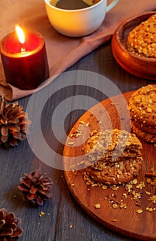 Sweet oatmeal cookies with cereals and black coffee in a white cup against the background of fir cones and a burning candle