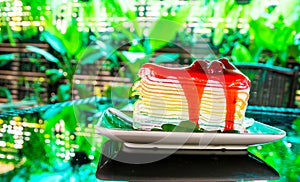 Sweet rainbow crape cake with strawberry sauce topping on plate on the table and shadow green tree background, snack menu at cafe.