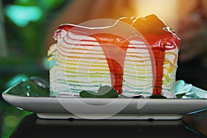 Sweet rainbow crape cake with strawberry sauce topping on plate on the table and shadow green tree background, snack menu at cafe.