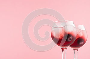 Sweet red alcoholic cocktails with ice cubes, blueberry in two goblets closeup, top section, edge in soft light pink background.