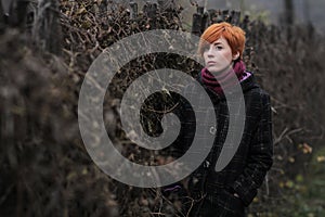 Sweet red-haired girl in a black coat and purple knitted scarf is standing by the fence overgrown with grapevine or ivy