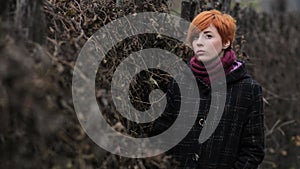 Sweet red-haired girl in a black coat and purple knitted scarf is standing by the fence overgrown with grapevine or ivy, girl or w