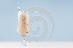 Sweet refreshing alcoholic creamy drink in goblet with ice cubes and straw in modern light blue interior on white wood table.