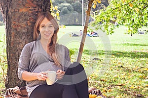 Sweet shot of attractive young woman expecting child sitting under tree, enjoying happy moment of her pregnancy, relaxing in open