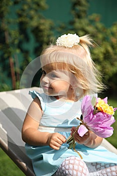 Sweet smiling little girl with long blond hair, sitting on grass in summer park, closeup outdoor portrait.