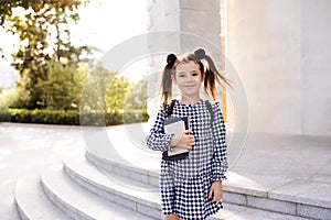 Sweet smiling young child girl 5-6 year old wear checkered black and white dress and backpack holding books stand outdoors close u