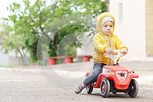 Sweet toddler boy in an orange jacket riding plastic toycar on the street in quiet neighborhood in the spring in cold photo