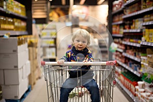 Sweet toddler boy, sitting in shopping cart, mother byuing groceries