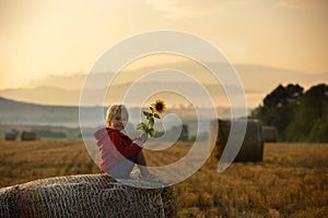 Sweet toddler child, boy, sitting on a haystack in field on sunrise