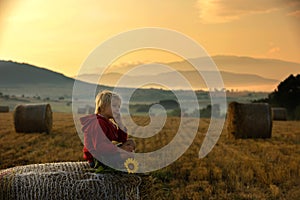 Sweet toddler child, boy, sitting on a haystack in field on sunrise