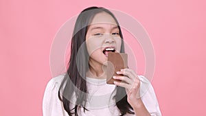 Sweet tooth. Studio portrait of little asian girl enjoying big chocolate bar and biting dessert, pink studio background
