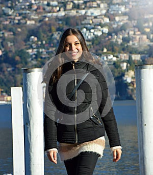 Sweet young woman with winter clothes walking on pier gangway of Lake Lugano with mountains on the background and flare sun light,