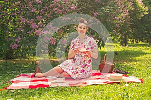 Sweetheart charming pinup girl in a summer dress on a checkered blanket in the Park near the bushes of lilac enjoys life and leisu