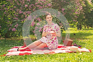 Sweetheart charming pinup girl in a summer dress on a checkered blanket in the Park near the bushes of lilac enjoys life and leisu