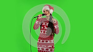 Sweety girl in Santa Claus hat is singing into a microphone and moving to the beat of music. Green screen
